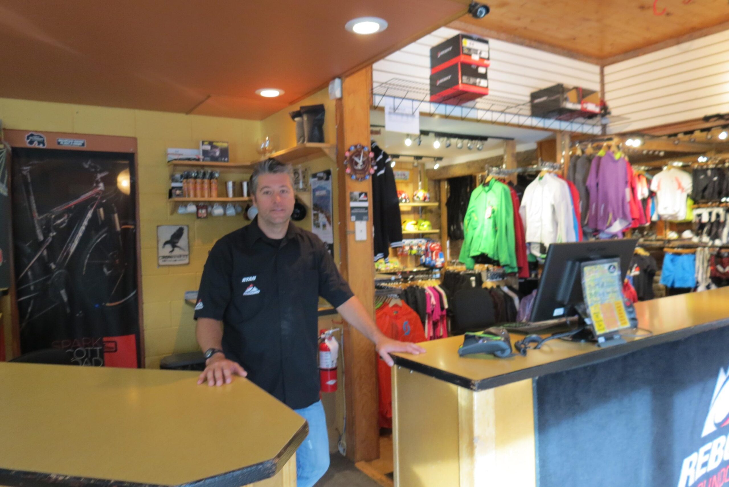 A man stands behind the counter of a retail store, with shelves displaying various outdoor clothing and gear in the background. The interior is well-lit, featuring wooden accents and colorful merchandise, including jackets and accessories. A computer and scanning equipment are visible on the counter.