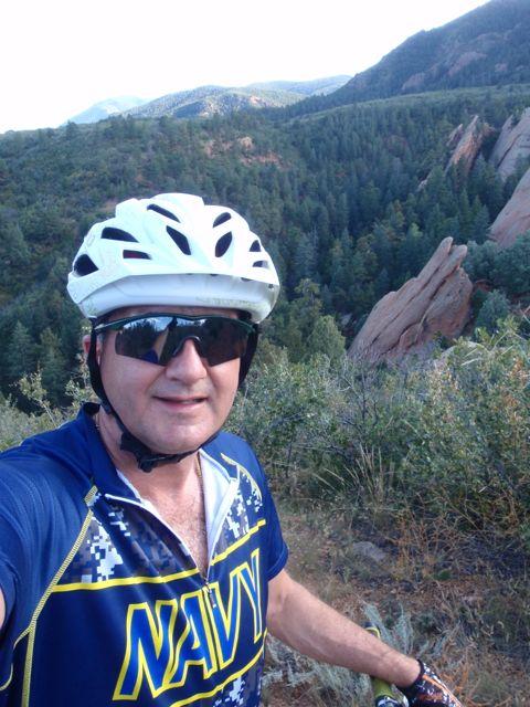 A person wearing a white helmet and sunglasses poses for a selfie while mountain biking on a scenic trail. They are dressed in a blue jersey with a yellow design, surrounded by lush green trees and rocky outcrops in the background. The landscape features rolling hills and a clear sky, indicating a beautiful outdoor setting. Red Rock Canyon mountain bike trail.