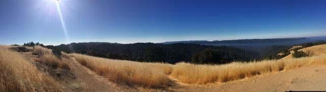 A panoramic view of rolling hills covered in tall, golden grass under a clear blue sky. The sun shines brightly in the upper left corner, casting light across a distant landscape of green trees and mountains. Saratoga Gap mountain bike trail.