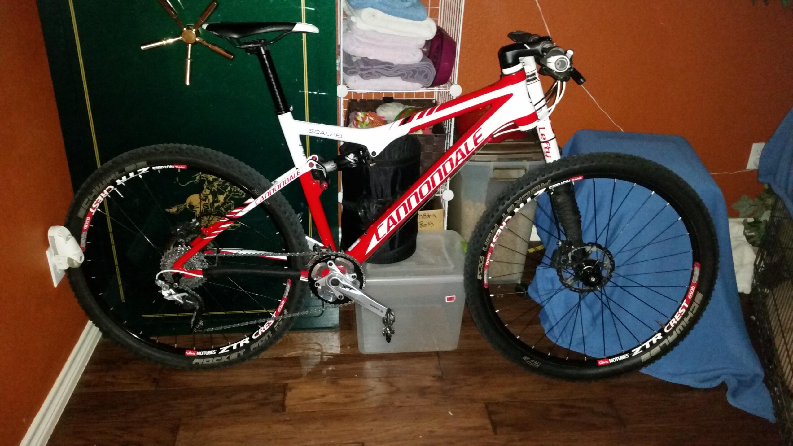 Cannondale Scalpel Carbon 3: A red and white Cannondale Scalpel mountain bike leaning against a wall, with one wheel mounted on a wall holder. In the background, there are towels on a rack and a storage container on the floor, with a blue blanket draped over a piece of furniture. The floor is wooden, and the room has an orange-painted wall.