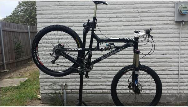 Santa Cruz Nomad: A mountain bike is positioned on a repair stand against a textured white wall, showcasing its black frame and wheels. The bike features prominent components like dual disc brakes and thick tires, indicating it's designed for off-road cycling. A small patch of grass and a fence are visible in the background.