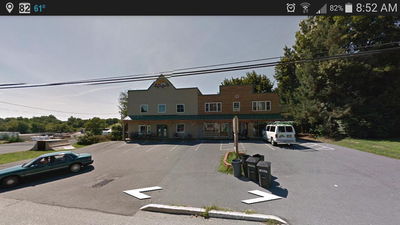 A parking lot in front of a two-story building with a mix of siding styles. The left side has a green awning with large windows, while the right side features a wooden facade. Several bicycles are parked near the entrance. A green sedan is positioned in the lot along with an older white van. In the foreground, there are three black garbage bins, and trees line the background.