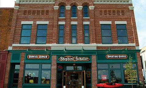 Historic brick building with large windows and a green storefront. Signs read "Service Repair" and "Shepherd Schaller Sporting Goods." A red kayak is displayed outside.