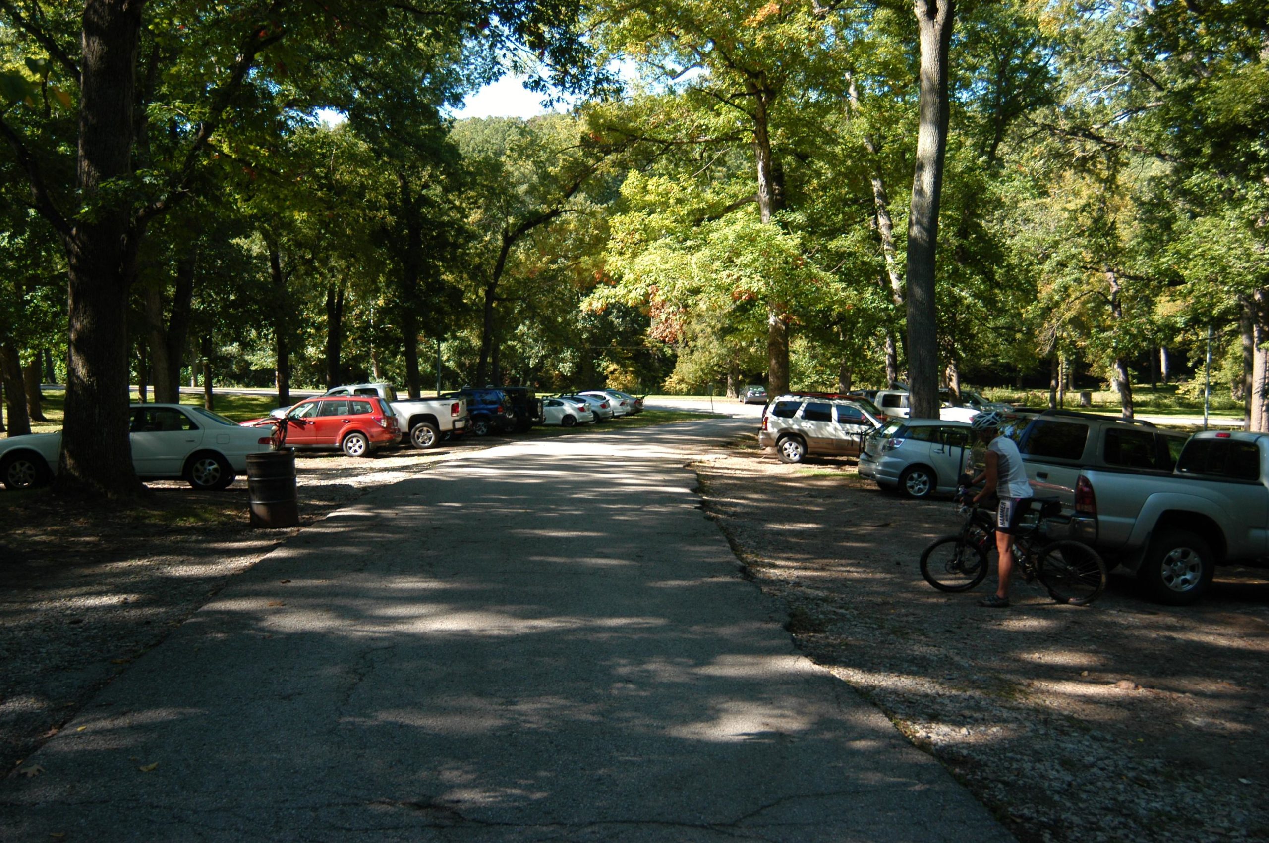 A wide view of a gravel parking area surrounded by trees, featuring several parked vehicles, including a red SUV and a white sedan. A cyclist in casual clothing stands next to a bicycle, taking a break at the side of the road, which is shaded by overhead trees. The scene is bright and sunny, indicating a pleasant day outdoors. Swope Park Trail mountain bike trail.