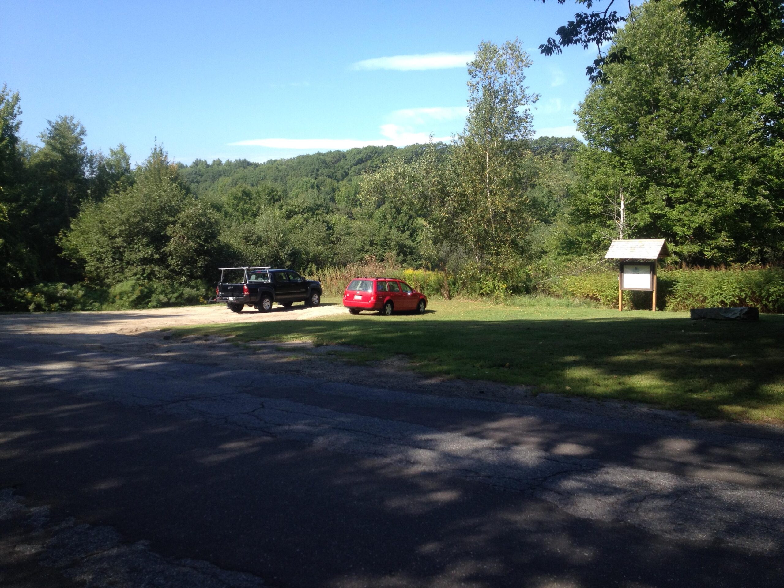 A scenic view of a parking area surrounded by trees and greenery, featuring a black vehicle and a red car parked on a gravel lot. To the right, there is an information board under a slanted roof. The background includes rolling hills under a clear blue sky. Tryon Mountain mountain bike trail.