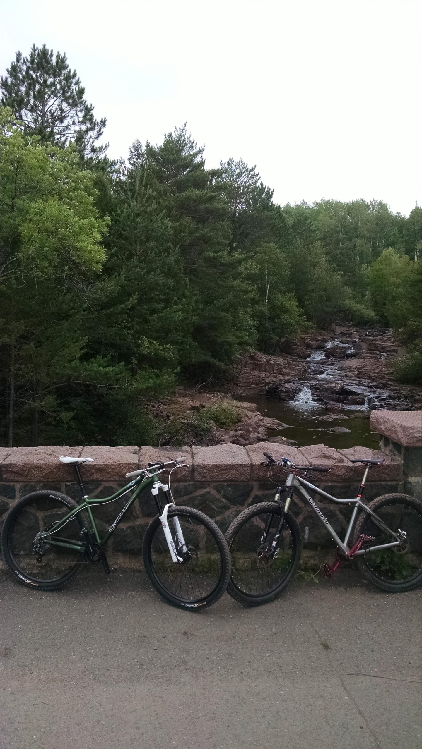 Canfield Brothers Yelli Screamy: Two mountain bikes, one green and black and the other silver and black, are resting against a stone wall overlooking a small rocky stream surrounded by lush greenery. The scene is set in a natural environment, with tall trees and a cloudy sky in the background.