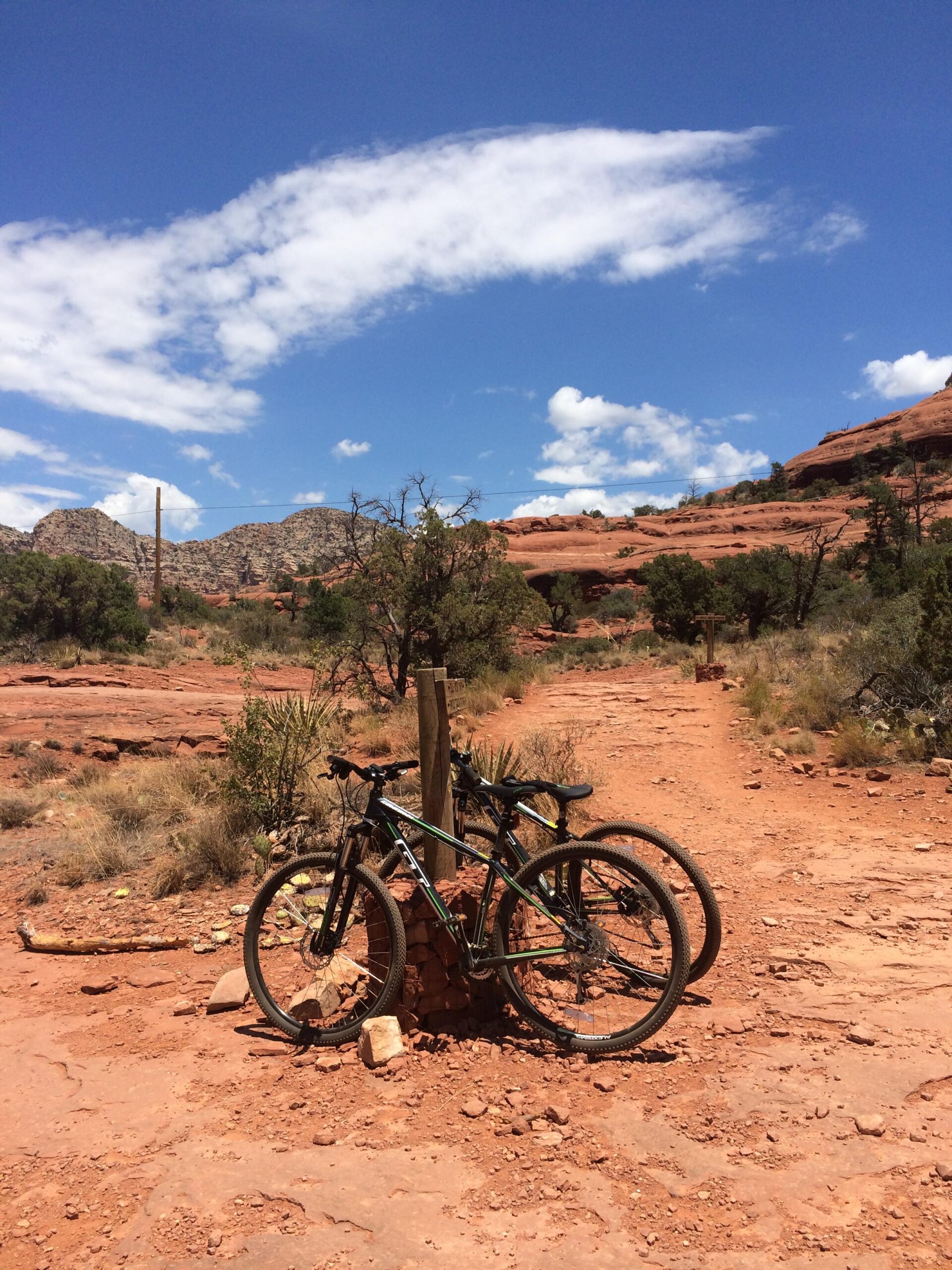GT Karakoram Sport 2014: Two mountain bikes are parked against a stone marker on a dirt trail surrounded by red rock formations and sparse vegetation. The scene is set under a clear blue sky with scattered clouds, showcasing a rugged, outdoor landscape ideal for biking and exploration.