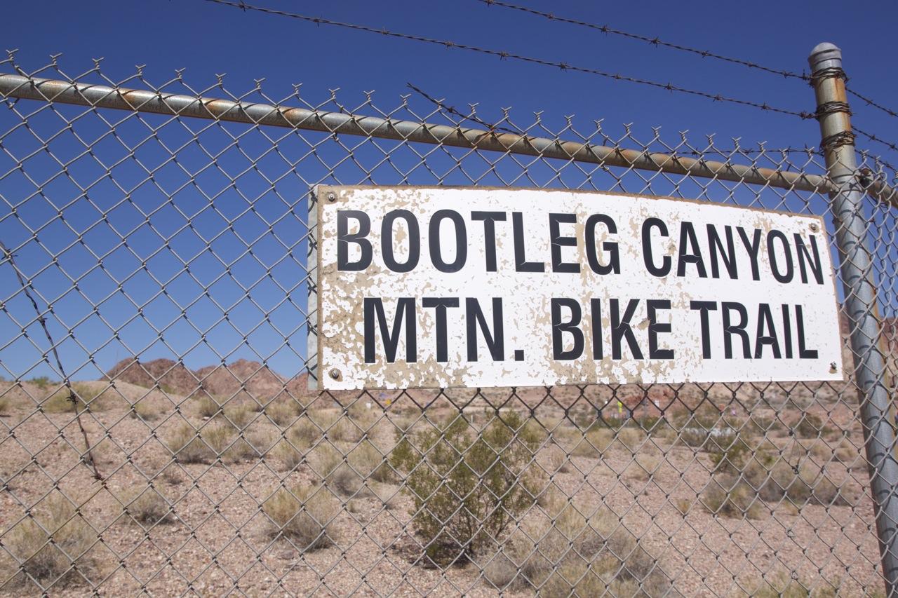 Sign for Bootleg Canyon Mountain Bike Trail, mounted on a chain-link fence, with a clear blue sky and rocky terrain in the background. The sign shows significant wear and tear, indicating it is well-used. Bootleg Canyon mountain bike trail.