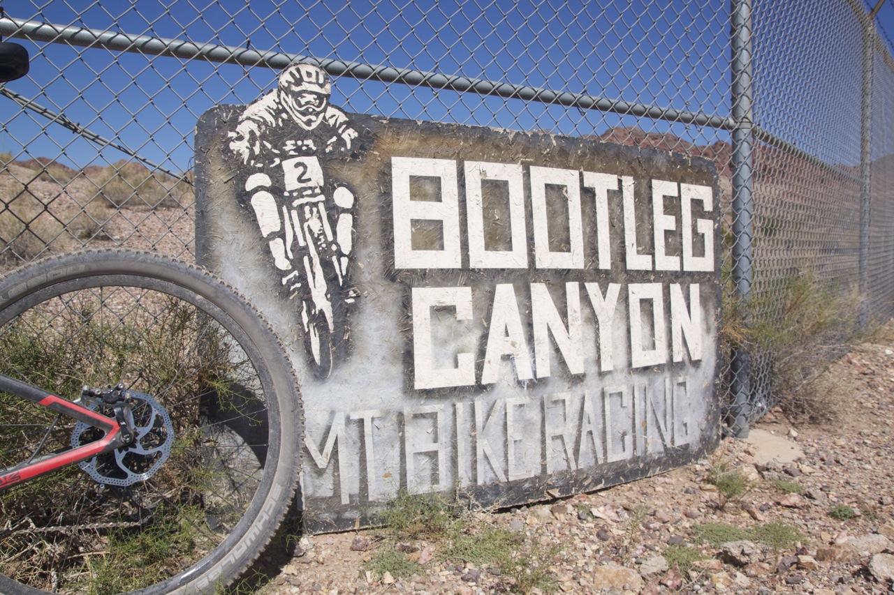 Sign for Bootleg Canyon Mountain Biking, featuring an illustration of a cyclist and text that reads "BOOTLEG CANYON MT BIKE RACING," with a mountain bike tire in the foreground and a chain-link fence in the background. Bootleg Canyon mountain bike trail.