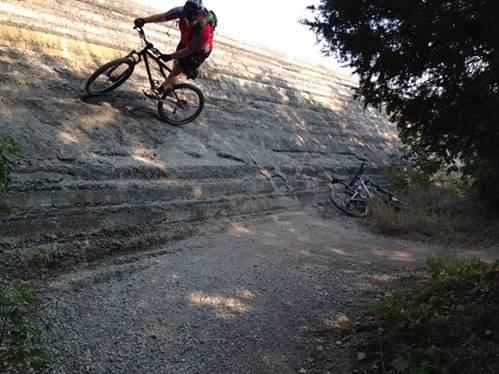 A mountain biker riding down a steep, gravel incline with another bicycle leaning against the slope in the background. The scene is surrounded by greenery and the texture of the ground is visible. Boulder Park mountain bike trail.