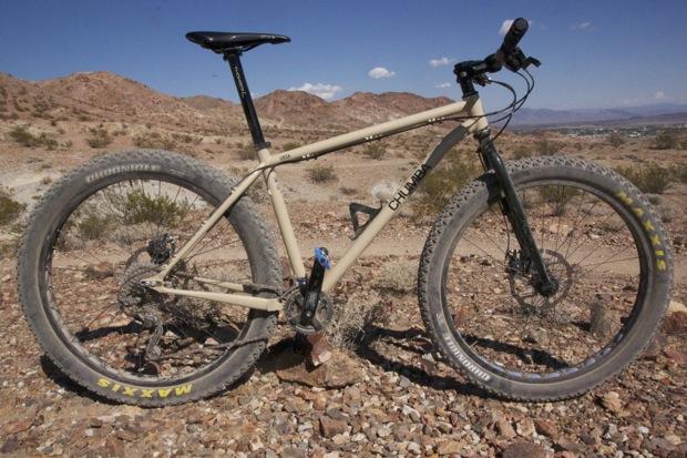 Chumba Ursa: A mountain bike placed on rocky terrain with a desert landscape in the background. The bike features a beige frame, large tires, and a front suspension, showcasing its suitability for off-road cycling. The scene includes hills and a clear blue sky.