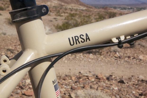Chumba Ursa: Close-up of a beige bicycle frame with the word "URSA" printed prominently on the top tube, featuring cables and a backdrop of rocky terrain and desert landscape.