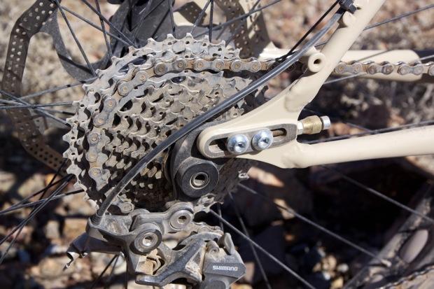 Chumba Ursa: Close-up of a bicycle's rear drivetrain components, including a cassette gear, chain, and derailleur, set against a gravel background.
