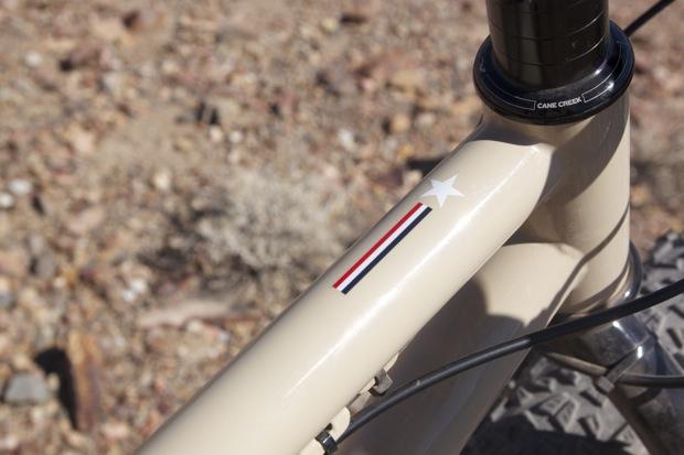 Chumba Ursa: Close-up of a beige bicycle frame featuring a decorative star and a stripe design in red, white, and blue, with rocky terrain in the background.