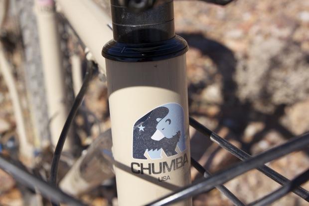 Chumba Ursa: A close-up view of a bicycle's frame featuring a "CHUMBA USA" logo, which includes a stylized bear graphic, set against a backdrop of rocky terrain. The frame is a light beige color with a black fork.
