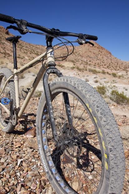 Chumba Ursa: A close-up view of a mountain bike in a rugged, rocky desert landscape. The bike features wide tires and a sturdy frame, with the handlebars in focus, set against a backdrop of dry, barren hills under a clear blue sky.