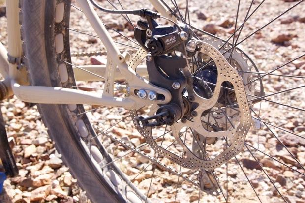 Chumba Ursa: Close-up view of a bicycle's rear disc brake assembly, featuring the brake rotor and caliper, set against a rocky ground background. The bike's frame is partially visible, showcasing a light-colored finish.