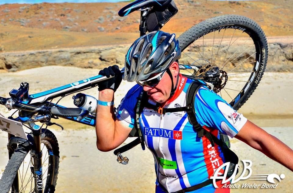 Trek Superfly 9.6: A cyclist wearing a blue and white jersey, helmet, and a backpack is carrying their mountain bike over a rocky terrain. The sun is shining brightly in a clear sky, highlighting the rugged landscape. The cyclist appears focused and determined as they navigate the challenging environment.