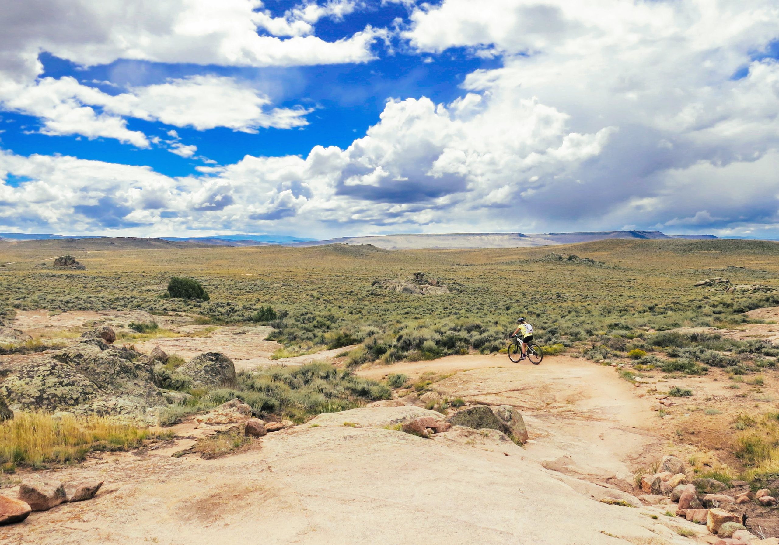A mountain biker rides along a rocky trail in a vast, open landscape under a blue sky dotted with fluffy white clouds. The area features patches of grass, sparse shrubs, and rugged terrain, highlighting the natural beauty of the outdoors. Hartman Rocks mountain bike trail.