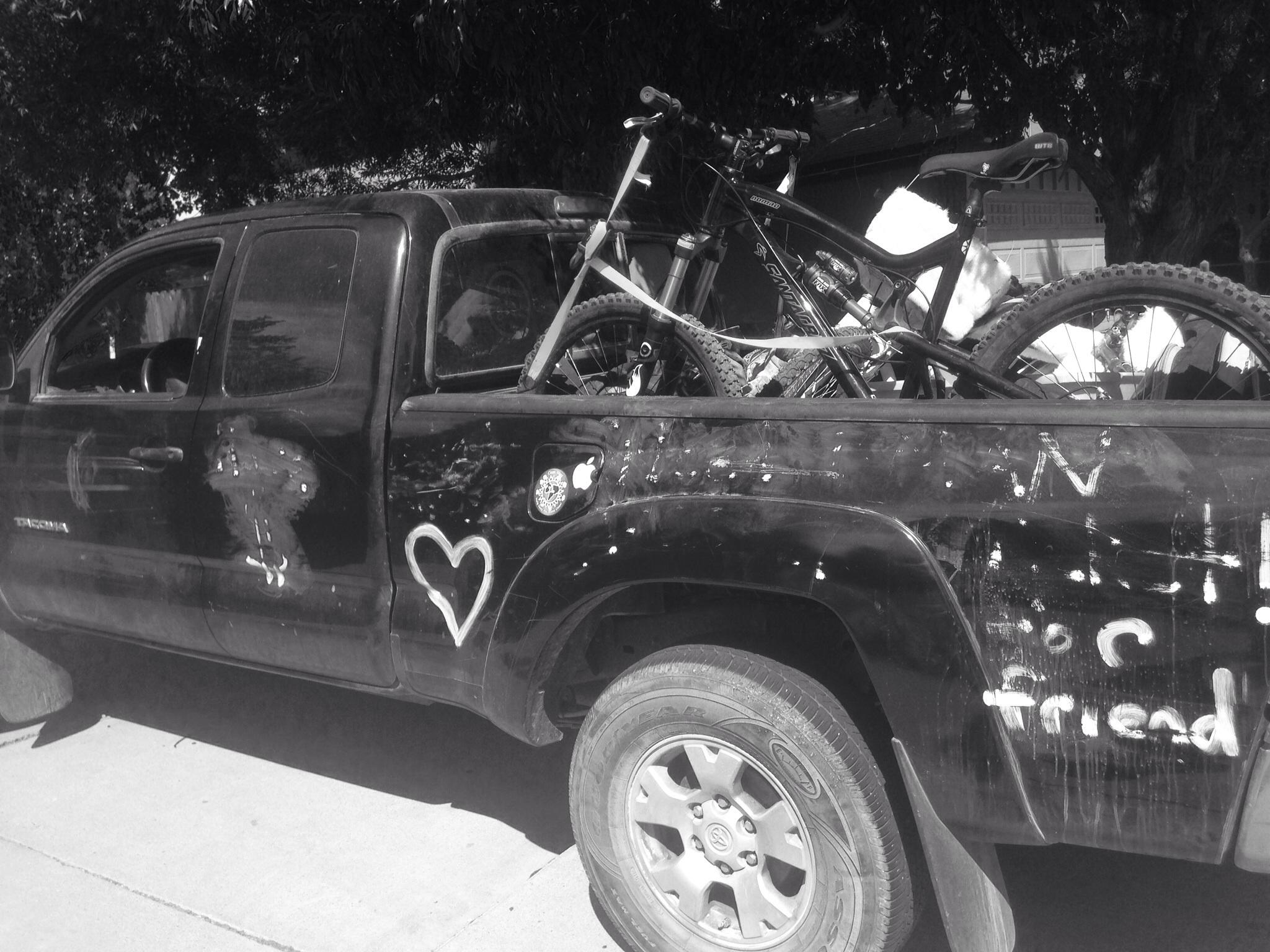 Santa Cruz Nomad: A black pickup truck parked with visible wear and tear, featuring a heart symbol and handwritten messages on its side. In the truck bed, two mountain bikes are visible, secured and ready for transport. The image is in black and white, with trees in the background partially shading the scene.