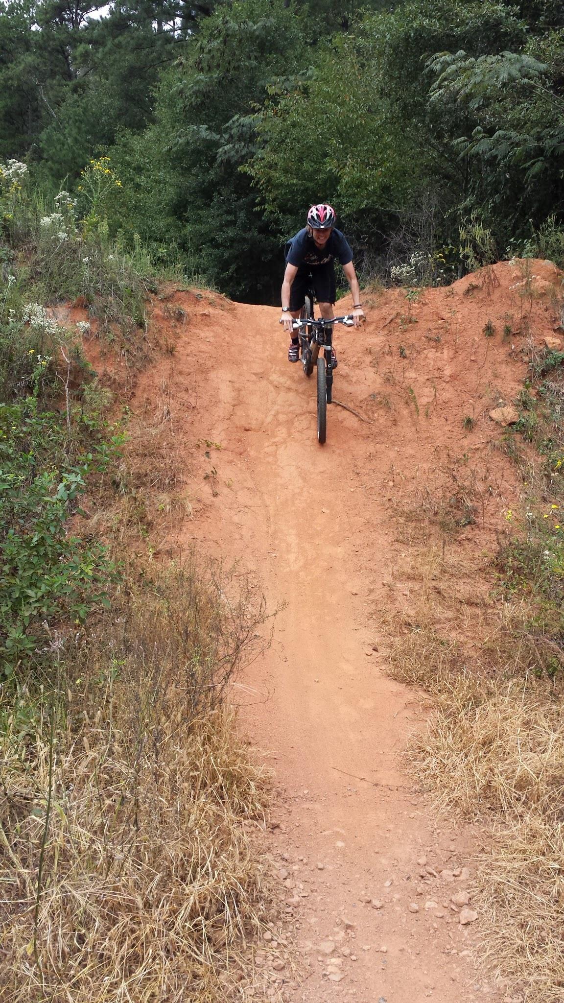 A mountain biker in a black shirt and helmet is descending a dirt ramp surrounded by greenery and shrubs. The biker is airborne, showcasing a dynamic leap off the ramp, with dirt and gravel visible on the trail below. Trail Creek Park mountain bike trail.