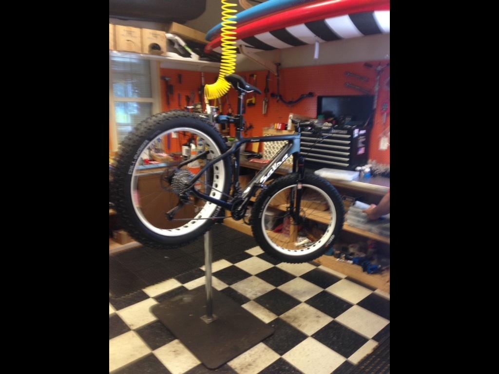 Salsa Beargrease: A fat bike mounted on a repair stand in a workshop, surrounded by tools and equipment. The floor features a black and white checkered pattern. Tools and parts are visible in the background, indicating a workspace for bicycle maintenance.