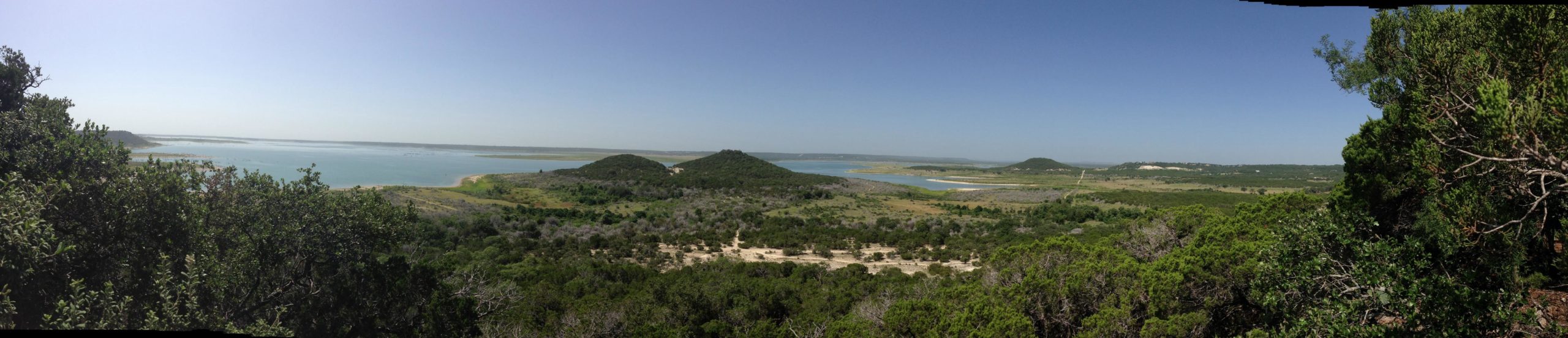 A panoramic view of a serene landscape featuring rolling hills, a blue lake, and lush green vegetation under a clear blue sky. The scene captures both water and land, showcasing the natural beauty of the area. Dana Peak mountain bike trail.
