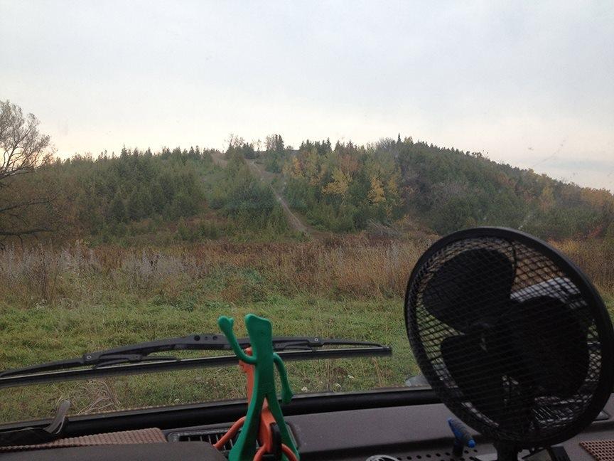 View from inside a vehicle looking out towards a hilly landscape covered with trees in various shades of green and yellow. In the foreground, a small fan is visible on the dashboard along with colorful clips. The sky is overcast, contributing to a calm, serene atmosphere. Harold Town Ca mountain bike trail.