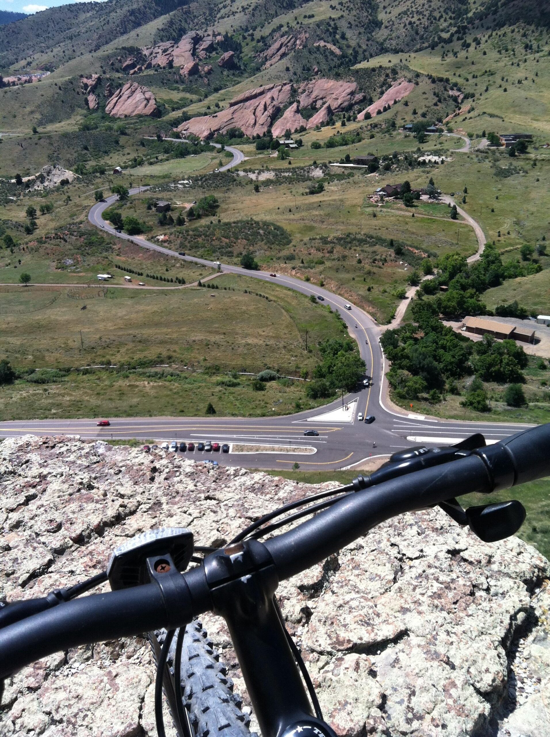 Trek 4500: A view from the handlebars of a mountain bike, overlooking a winding road and green landscape. In the distance, large rock formations and hills are visible under a clear blue sky. The scene showcases the natural beauty of the area, with trees and grassy fields surrounding the road below.