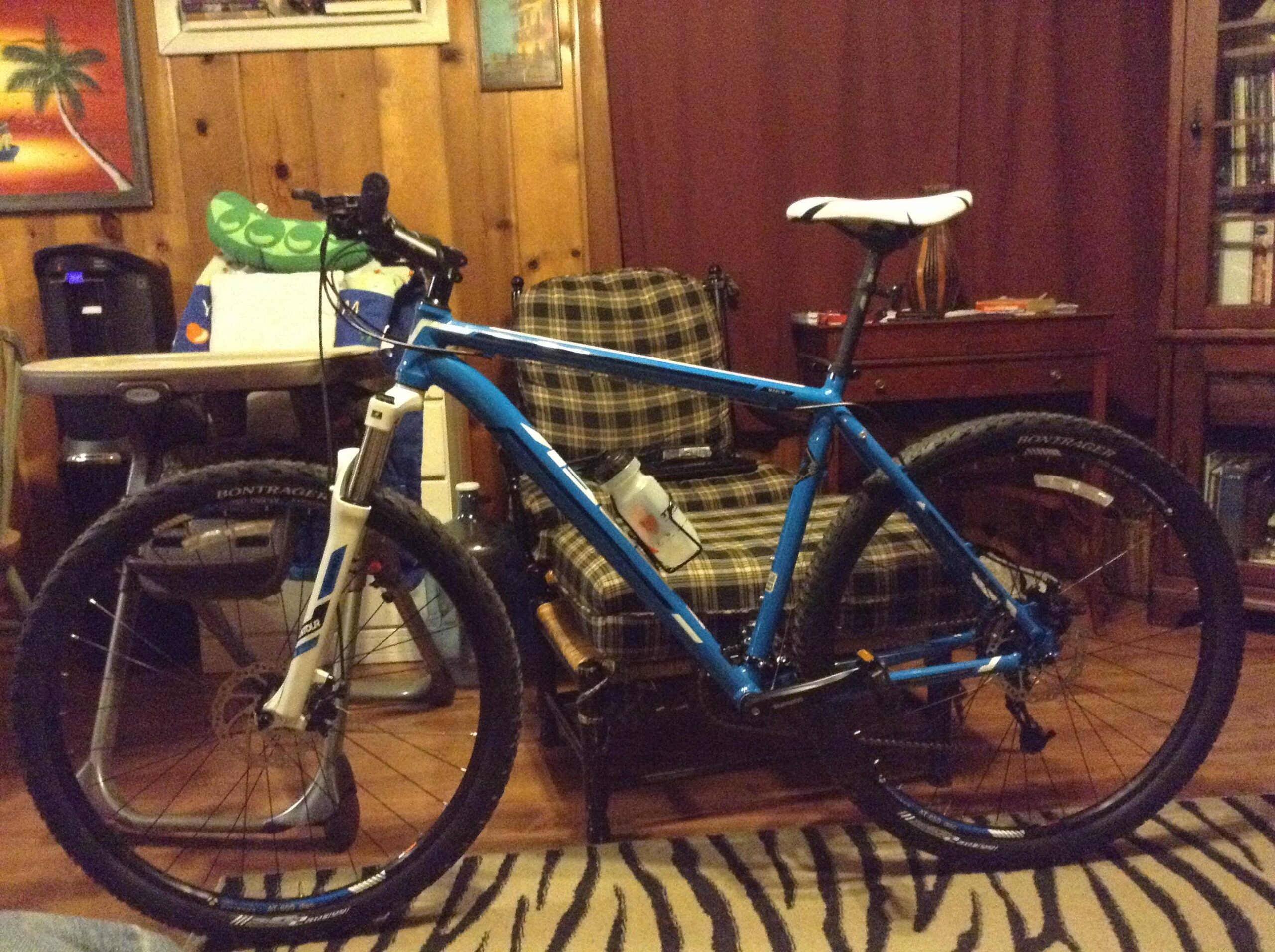 Trek Marlin: A blue mountain bike is leaning against a wooden chair in a rustic room with wooden walls. The bike features a white seat and black tires, with a water bottle holder attached to the frame. In the background, there is a plaid chair, a table with various items, and a bookshelf. The floor has a zebra-striped rug.
