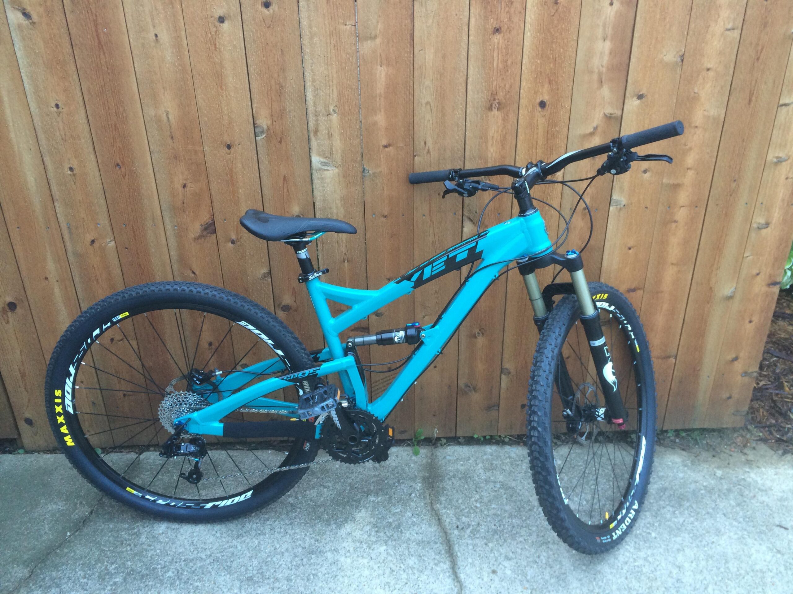 Yeti SB95: A vibrant turquoise mountain bike leaning against a wooden fence, featuring a sleek frame, black tires with white accents, and a comfortable saddle. The bike's components, including the handlebars and gears, are clearly visible, showcasing its design for off-road riding.