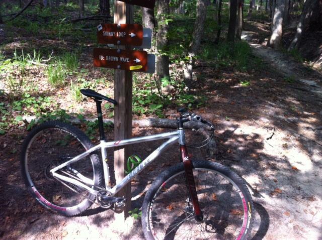 Niner One 9: A white mountain bike resting against a wooden trail sign in a wooded area. The sign points to two trails: "Skinny Loop" and "Brown Wave." The ground is covered in dirt and leaves, with sunlight filtering through the trees in the background.
