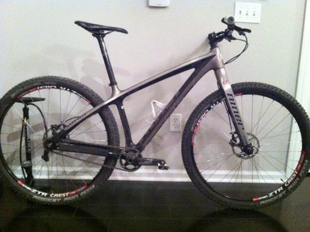 Niner Air 9 Carbon: A modern mountain bike with a lightweight carbon frame, featuring oversized tires for traction. The bike has disc brakes, a sleek design, and is set against a neutral wall. A bike pump is leaning next to it, and there are visible brand logos on the rims.