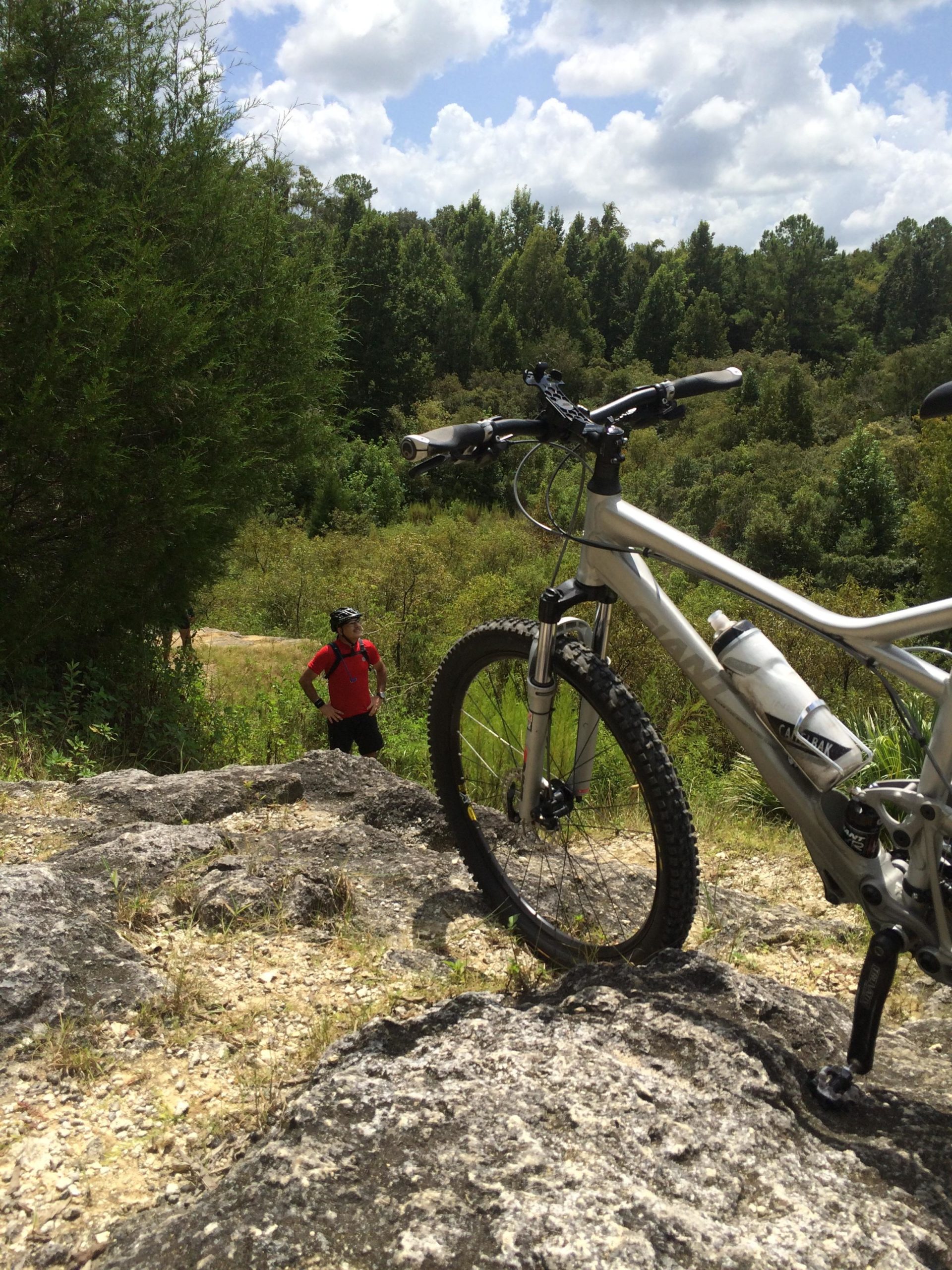A mountain bike leaning against a rocky outcrop, with a lush green forest in the background and a person in a red shirt standing nearby on a trail. The sky is partly cloudy, creating a bright outdoor scene. Santos mountain bike trail.