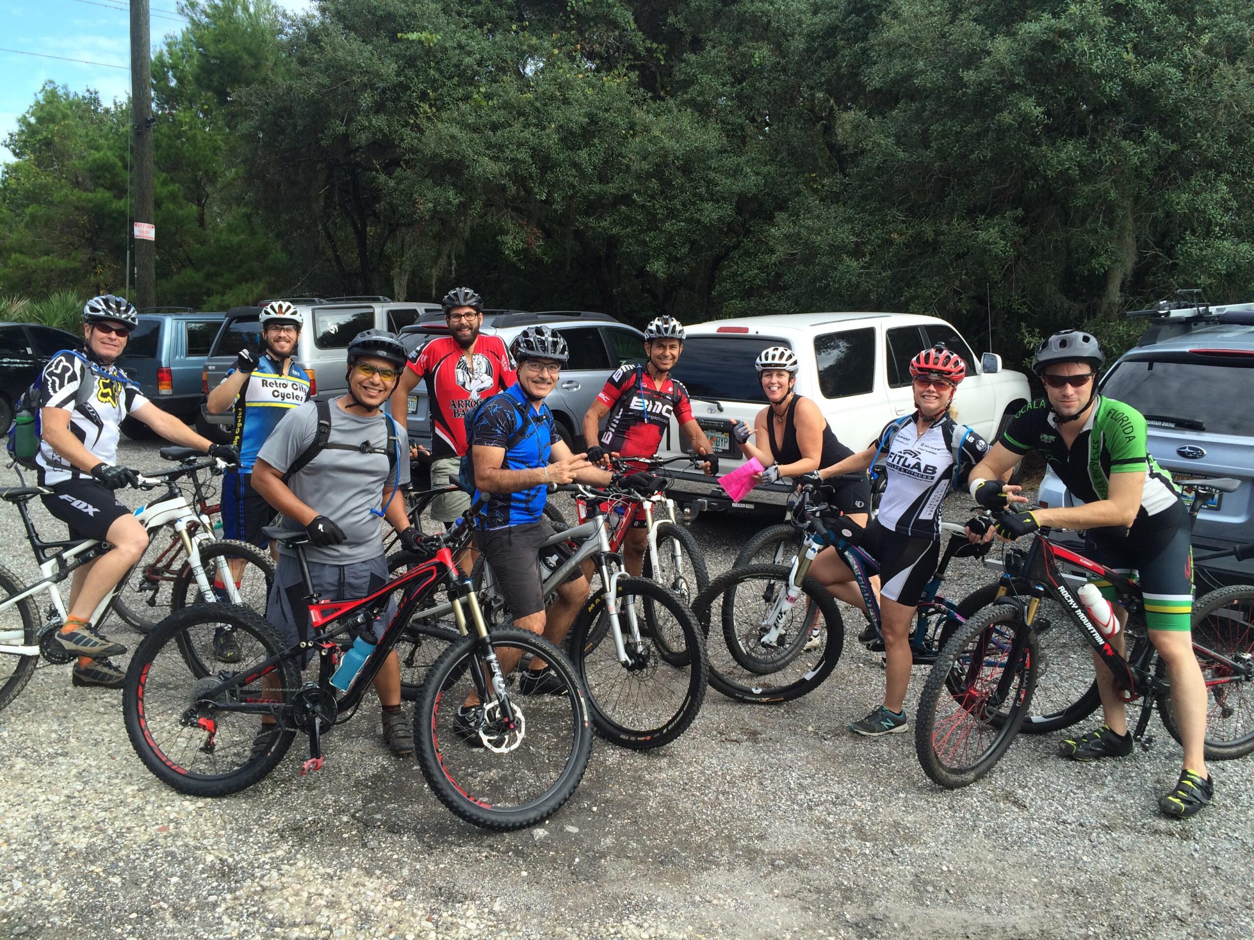 A group of eight cyclists posing for a photo, each wearing cycling gear and helmets while standing next to their mountain bikes. They are gathered in a parking area surrounded by trees and parked vehicles, smiling and appearing ready for a ride. Balm Boyette Scrub Preserve mountain bike trail.