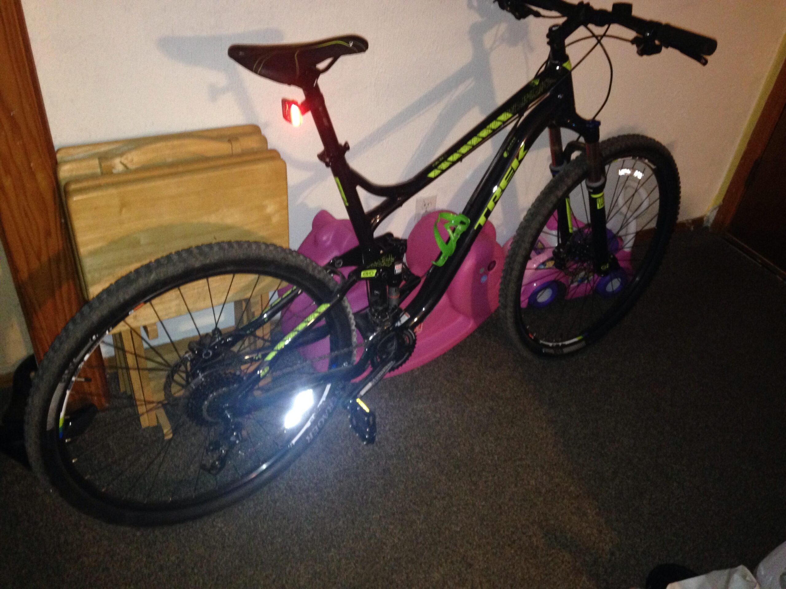 Trek Fuel EX 5 29: Alt text: A black mountain bike with green accents is leaning against a wall in a room. The bike features a rear red light and has a reflective surface on one of the wheels. Nearby, there are two folded wooden tables and a pink object, possibly a child's toy. The floor is carpeted.
