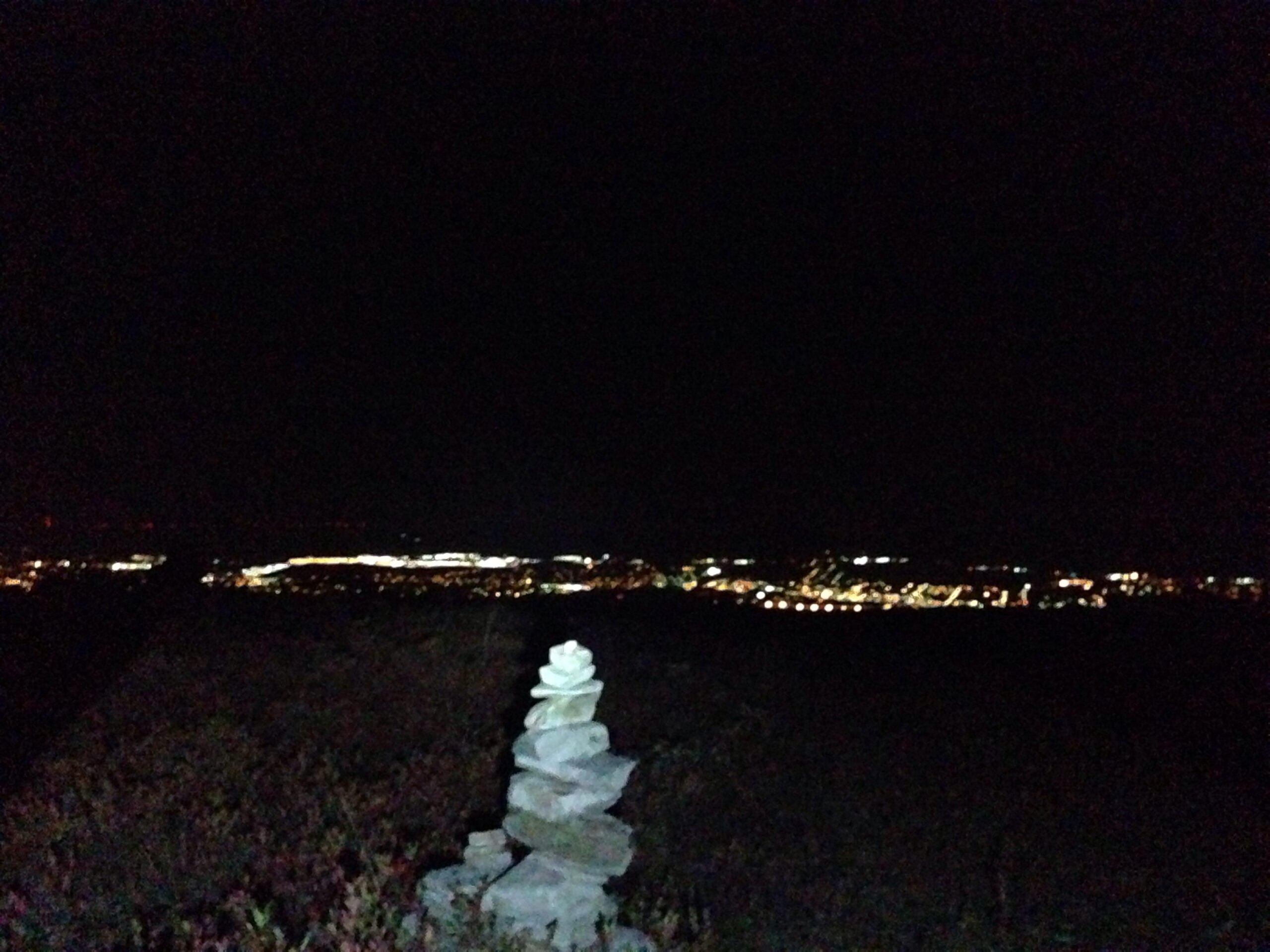 A stack of stones is prominently displayed in the foreground, with a dark night sky above. In the background, faint lights from a distant city twinkle against the dark landscape. The image conveys a peaceful nighttime scene with natural elements juxtaposed against urban lighting. Blueberry mountain bike trail.