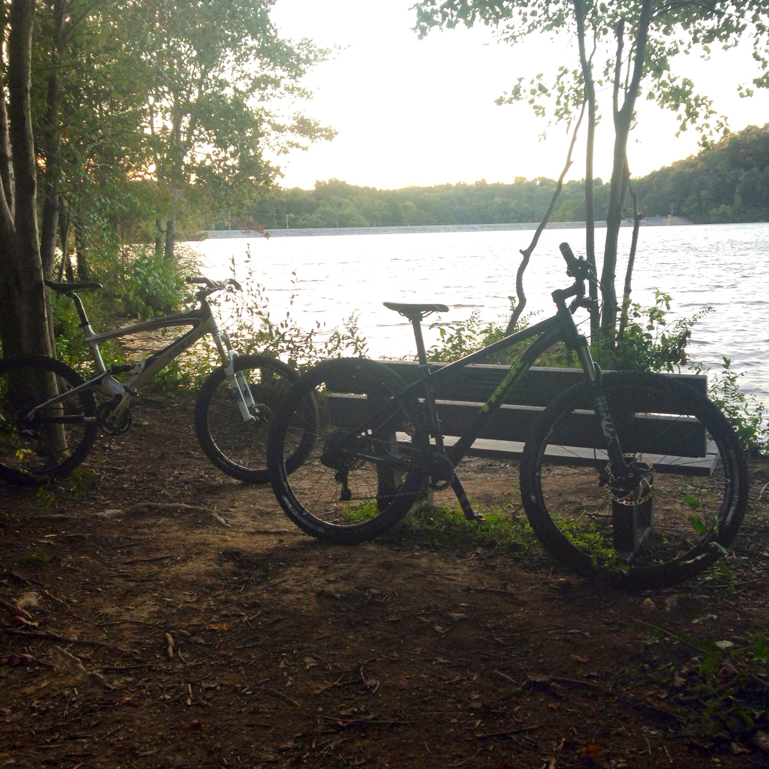 Trek Stache 8: Two mountain bikes parked beside a tranquil lake at sunset, surrounded by trees and greenery. A wooden bench is visible nearby, with gentle ripples on the water reflecting the soft light. The scene conveys a peaceful outdoor atmosphere ideal for cycling and nature appreciation.