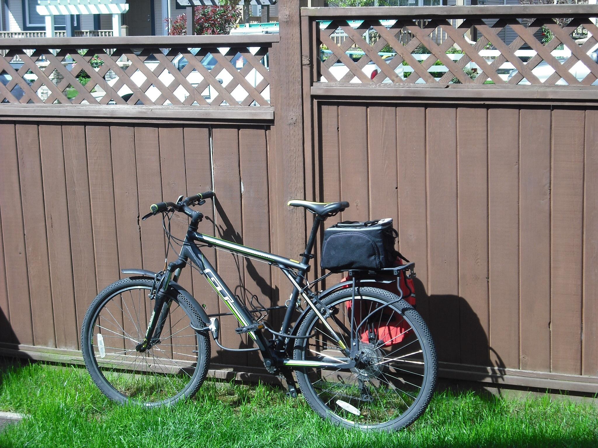 GT Aggressor 3.0: A black and yellow mountain bike resting against a wooden fence, with a green patch of grass in the foreground. The bike is equipped with a black bag attached to the rear. The fence features a lattice design at the top and parts of houses and greenery are visible in the background.