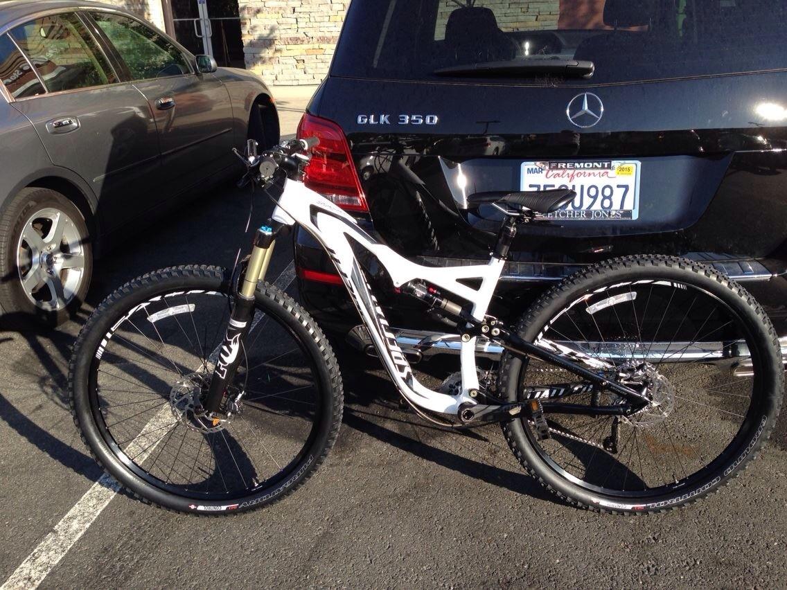 Specialized Stumpjumper FSR Elite: A mountain bike with a white and black frame is parked beside a black Mercedes GLK 350 in a parking lot. The bike features thick tires and a front suspension fork. The background includes a gray car and a building with a stone facade.