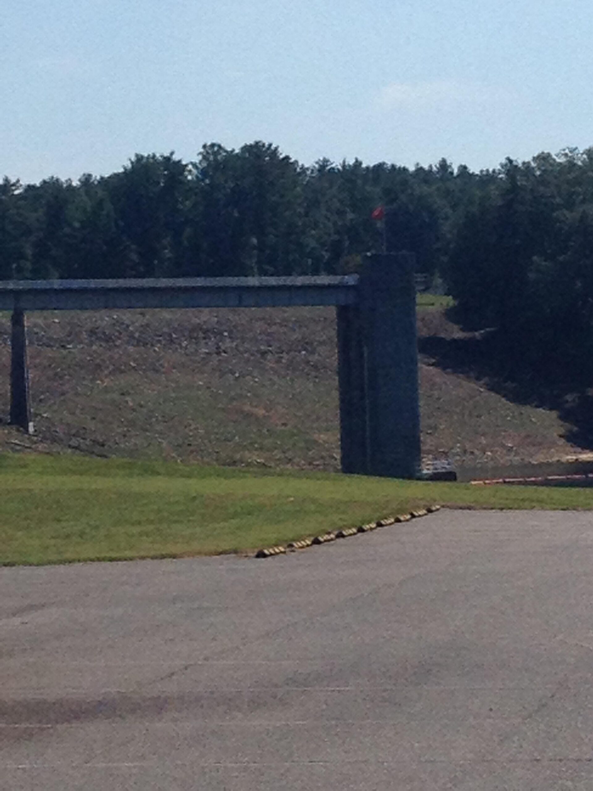 A concrete bridge spans a low reservoir area, with a flag on one of the supporting structures. In the background, a dense line of trees is visible under a clear sky. The foreground features a paved area, suggesting a parking lot. Dark Mountain Trail mountain bike trail.