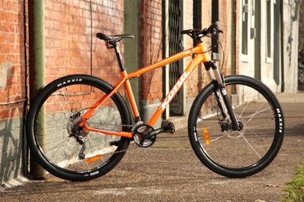 Kona Kahuna: An orange mountain bike rests against a textured brick wall in an urban setting. The bike features sturdy Maxxis tires and is positioned on a paved surface, with windows and doors visible in the background. The overall scene conveys a vibrant, adventurous atmosphere.
