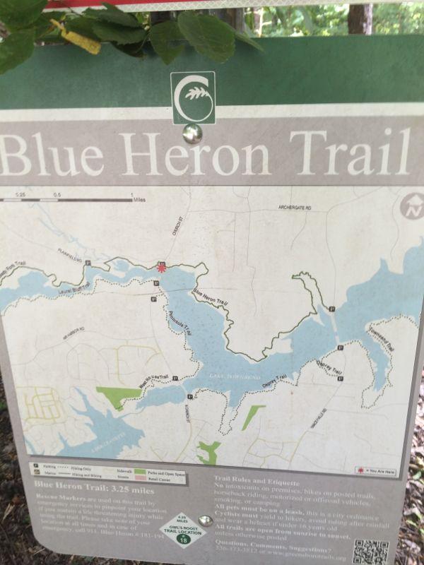 Map of the Blue Heron Trail, which is 3.25 miles long, displayed on a trail sign. The map shows the trail's path, surrounding water bodies, and nearby roads. There are notes on trail rules and etiquette at the bottom of the sign. The sign is partially obscured by leaves. Blue Heron mountain bike trail.
