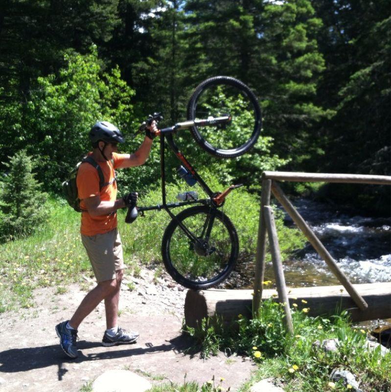 A cyclist wearing a helmet and orange shirt lifts a mountain bike above his head while standing on a dirt path near a stream. Lush green trees and grass surround the area, creating a natural outdoor setting. South Cottonwood mountain bike trail.