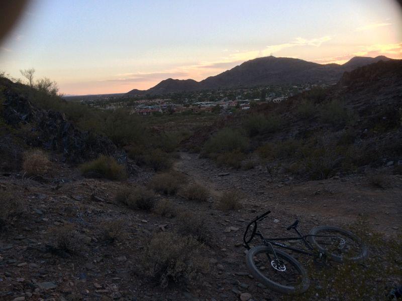 A rocky trail in a desert landscape at sunset, with a mountain range in the background and a city visible in the distance. A mountain bike is resting on the ground, surrounded by sparse vegetation and shrubs. The sky features soft hues of orange and purple as the sun sets behind the mountains. Trail #100 mountain bike trail.