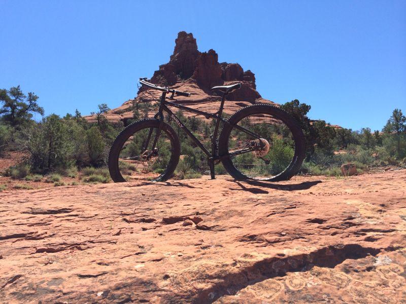 A black mountain bike is positioned on a rocky terrain in a desert landscape, with a prominent red rock formation rising in the background under a clear blue sky. Bell Rock Trailway mountain bike trail.