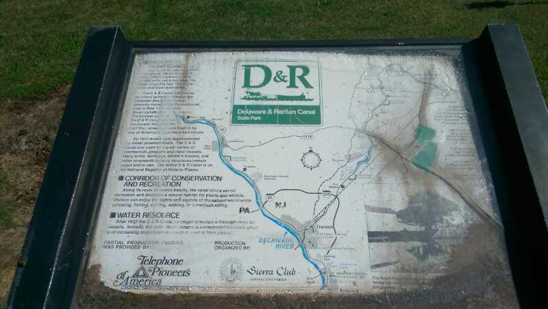 A weathered informational sign for the Delaware and Raritan Canal State Park, featuring a map of the park and surrounding areas, with descriptions related to conservation, recreation, and water resources. Six Mile Run mountain bike trail.