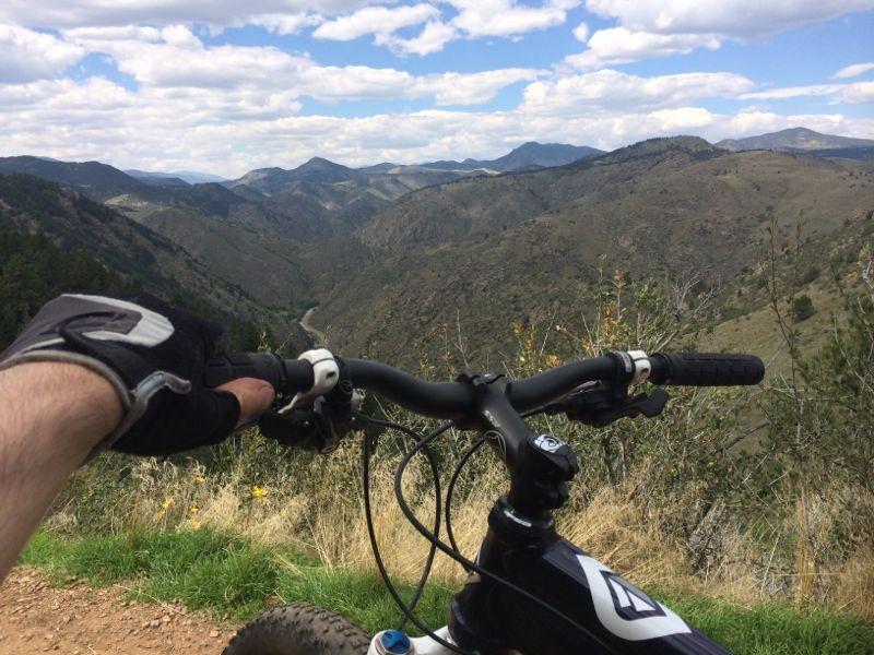 A mountain biker's perspective from the handlebars, overlooking a scenic valley with rolling hills and mountains in the background. The sky is partly cloudy, and greenery is visible in the foreground. Chimney Gulch mountain bike trail.