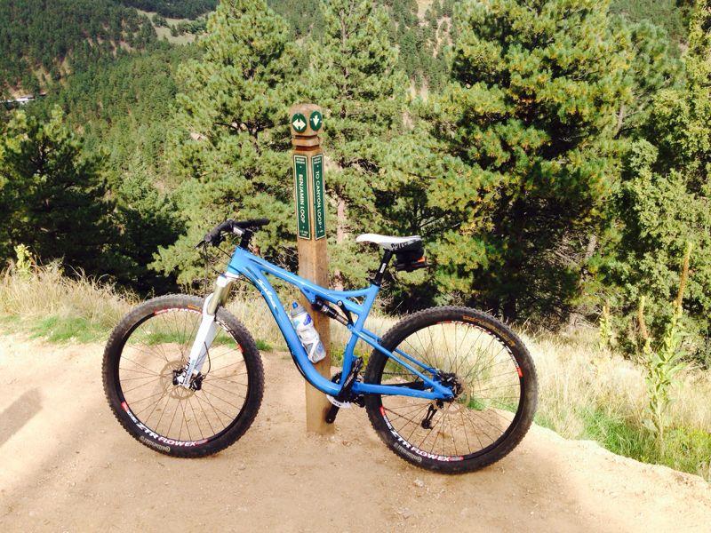 A blue mountain bike parked next to a trail marker in a lush, green landscape, surrounded by pine trees and a dirt path. The marker indicates trail directions for hikers and bikers. Betasso Preserve mountain bike trail.