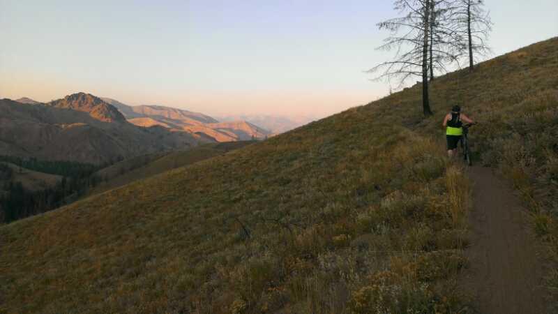A cyclist walking their bike along a dirt trail on a sloped hillside, with mountains in the background. The scene is illuminated by the warm light of sunset, showcasing the colorful landscape of grass and rocky terrain. Sparse trees are visible in the foreground, and distant mountains create a hazy backdrop. Imperial Gulch mountain bike trail.